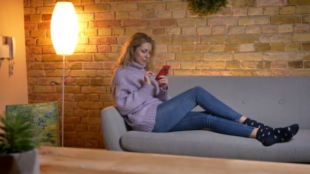 Nahaufnahme Shooting der erwachsenen kaukasischen Blondine mit dem Telefon, während sie entspannt auf der Couch sitzt und ihre Beine in ihrem gemütlichen Zuhause spreizt — Stockvideo