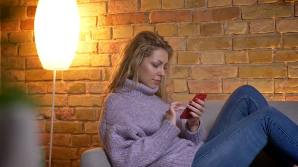 Närbild skjuta av vuxen kaukasiska blond kvinna surfar på telefonen medan du sitter på den soffan laidback vilar inomhus för mysiga hemma — Stockvideo