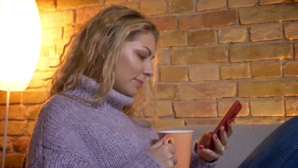 Gros plan côté vue shoot de adulte caucasien blonde femme navigation sur le téléphone tandis que assis sur le canapé décontracté et boire du café chaud — Video