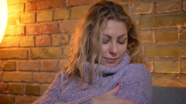 Closeup shoot of adult caucasian blonde female warming herself in a cozy warm sweater while sitting on the couch indoors at cozy home — Stock Video