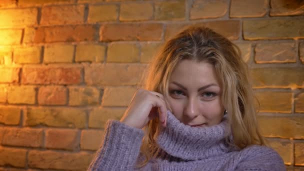 Closeup shoot of adult caucasian blonde female hiding her cute face in a warm sweater and looking at camera with joy while sitting on the couch indoors at cozy home — Stock Video