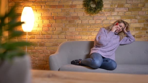 실내에 아늑한 집에서 소파에 앉아있는 동안 전화에 캐주얼 대화를 나누는 성인 백인 금발 여자의 근접 촬영 — 비디오