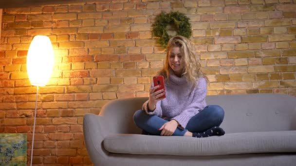 Closeup shoot of adult caucasian attractive female having a video call on the phone while sitting on the sofa indoors in a cozy apartment — Stock Video