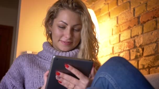 Closeup přední střílet z dospělých kavkazské blondýna ženské textilie na tabletu, při pohledu na fotoaparát a vesele zatímco sedí na gauči doma v útulném bytě — Stock video