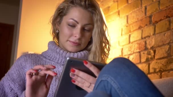Крупним планом передня зйомка дорослих білошкірих жіночих смс на планшеті, сидячи на дивані в приміщенні в затишній квартирі — стокове відео