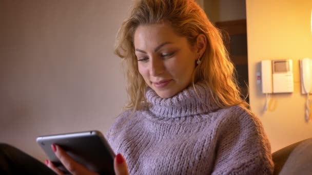 Primer plano de rodaje de adulto caucásico rubia hembra navegando en la tableta y sonriendo alegremente mirando a la cámara interior en un acogedor apartamento — Vídeos de Stock