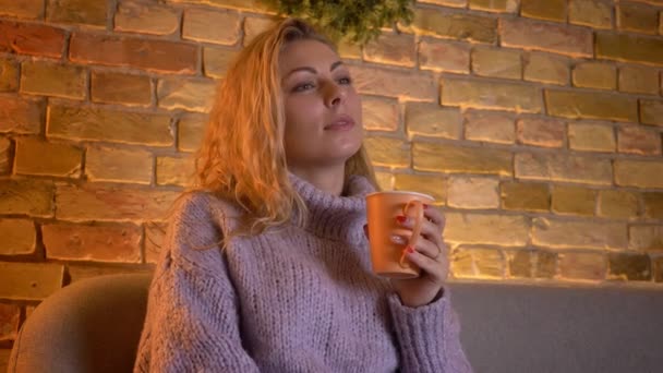 Крупним планом знімок дорослої білої жінки, яка дивиться телевізійне комедійне шоу, що тримає чашку теплого чаю, сидячи на дивані в приміщенні в затишному будинку — стокове відео