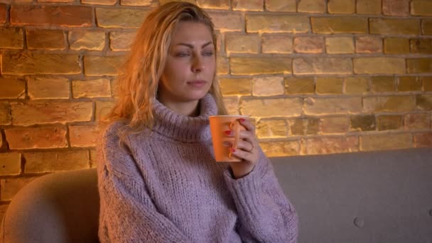 Closeup shoot of adult caucasian blonde female watching TV with curious facial expression holding a cup of warm tea while sitting on the couch indoors at cozy home — Stock Video