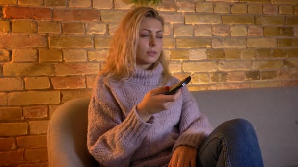 Vértes lő felnőtt kaukázusi szőke női néz Tv-val kíváncsi arckifejezés és a távirányító használata közben ül a kanapén bent a meghitt otthon — Stock videók