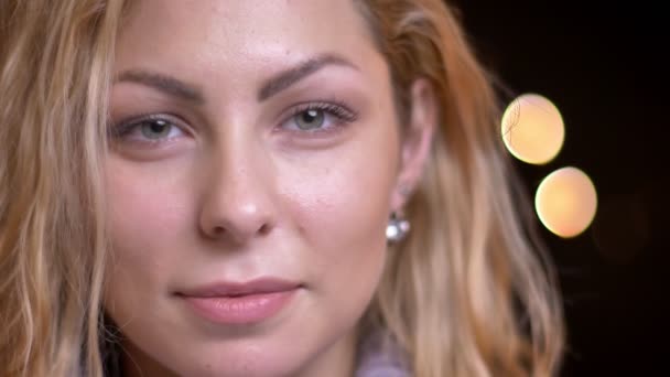 Nahaufnahme einer erwachsenen attraktiven blonden Kaukasierin, die direkt in die Kamera schaut und fröhlich mit Bokeh-Lichtern auf dem Hintergrund lächelt — Stockvideo