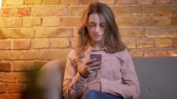 Porträtt av unga attraktiva tonårs flicka sitter på soffan och tittar in i smartphone i mysig hem atmosfär. — Stockvideo