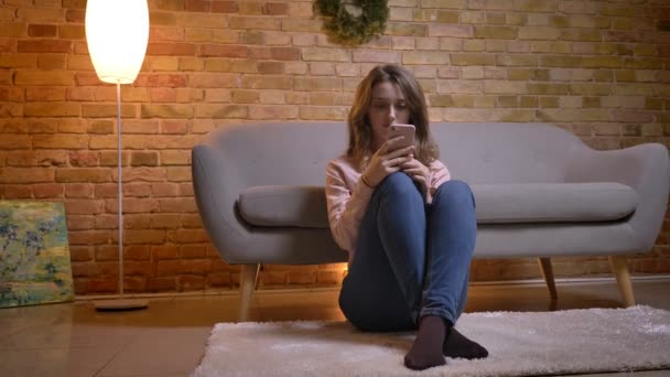 Retrato de una joven estudiante sentada en el suelo con las rodillas dobladas mirando hacia el teléfono celular con atención en un acogedor ambiente hogareño . — Vídeos de Stock