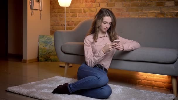 Retrato de una joven estudiante sentada en el suelo mirando hacia el teléfono celular y reaccionando tranquilamente en un ambiente acogedor en casa . — Vídeos de Stock