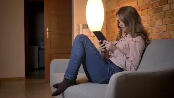 Portrait in profile of young teenage girl watching attentively into tablet and adjusting her curly hair in cozy home atmosphere. — Stock Video