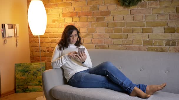 Porträtt av attraktiv brunett affärs kvinna som ligger på soffan och tittar uppmärksamt i smartphone hemma. — Stockvideo
