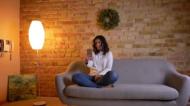 Beyaz esmer ev hanımı kanepe üzerinde oturan ve evde akıllı telefonda videochat konuşurken portresi.
