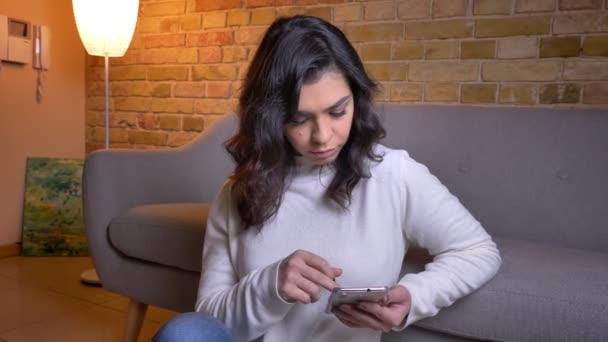 Gros plan portrait de belle femme d'affaires brune caucasienne assise sur le sol et regardant attentivement dans le smartphone à la maison . — Video