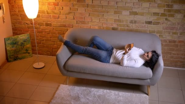 Atractiva mujer de negocios morena relajándose en el sofá y viendo en el teléfono inteligente en casa . — Vídeo de stock