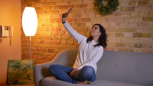 Hermosa morena caucásica ama de casa sentada en el sofá y haciendo selfie-fotos usando el teléfono inteligente alegremente en casa . — Vídeos de Stock