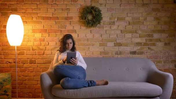 Портрет привабливої брюнетки домогосподарка сидить на дивані і радісно дивиться в планшет вдома . — стокове відео