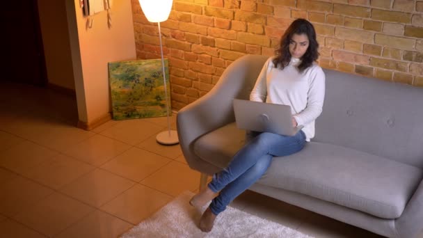 Κορυφαία βολή της προσεκτικής καυκάσιας μελαχρινή επιχειρηματίας κάθεται στον καναπέ με το laptop αλλάζει τη θέση των ποδιών στο σπίτι. — Αρχείο Βίντεο
