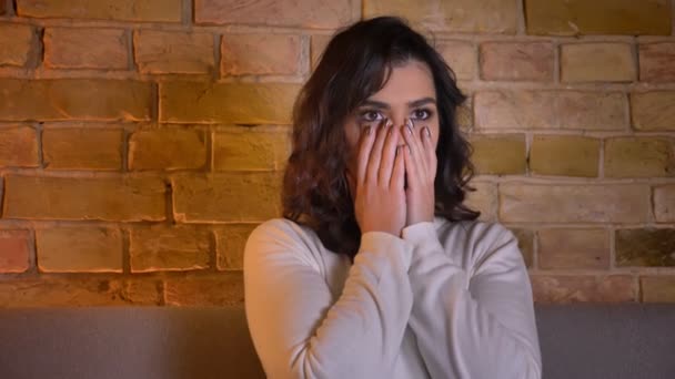 Scared caucasian brunette woman watching horror movie and covering her face in fear in cozy home atmosphere. — Stock Video