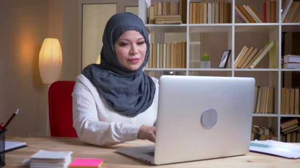 Close-up shoot van volwassen moslim succesvolle zakenvrouw te typen op de laptop te kijken naar de camera en glimlachend gelukkig op de werkplek binnenshuis — Stockvideo