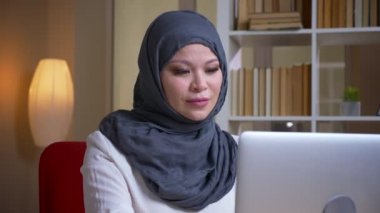 Yetişkin Müslüman başarılı iş kadını kamera bakarak laptop yazarak ve işyerinde kapalı gülümseyen çekim çekim