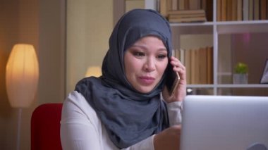 İş yerinde kapalı dizüstü bilgisayar kullanırken telefon üzerinde resmi bir konuşma sahip hijab yetişkin Müslüman işkadını çekim çekim