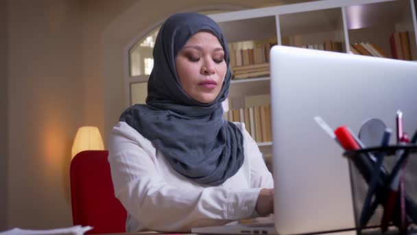 Zbliżenie strzelać dorosłych muzułmańskich udane businesswoman wpisując na laptopie i patrząc na aparat uśmiechnięty szczęśliwie na miejscu pracy w pomieszczeniu — Wideo stockowe
