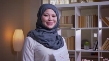 Kütüphanedeki grafik okuyan hijab yetişkin Müslüman kadın çalışanın closeup ateş düşünceli ve kamera neşeli işyerinde kapalı gülümsüyor bakıyor