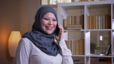 Hijab içinde çalışma alanında kütüphanede telefonda konuşan neşeli yetişkin Müslüman işkadını çekim çekim kapalı