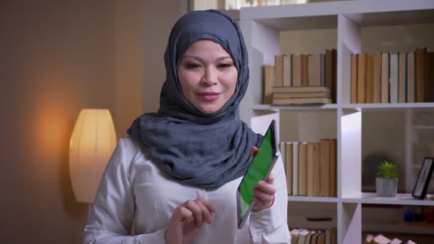 Nærbilde av en voksen muslimsk forretningskvinne som bruker nettbrettet med grønn skjerm foran kamera mens hun smiler lykkelig på arbeidsplassen innendørs – stockvideo