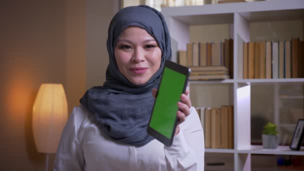 Closeup shoot of adult muslim female employee in hijab using the tablet and showing green chroma screen to camera smiling happily on the workplace indoors — Stock Video