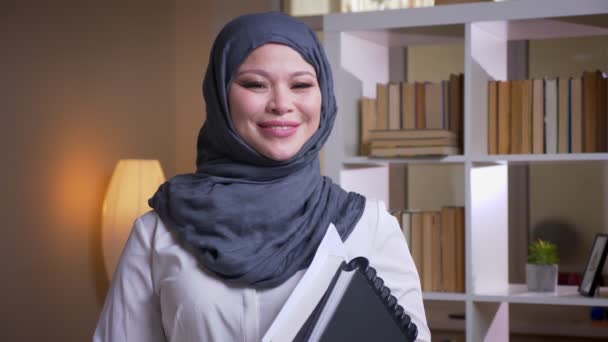 特写镜头拍摄的成年穆斯林女医生在头巾拿着书, 直视相机愉快地微笑在图书馆在室内 — 图库视频影像
