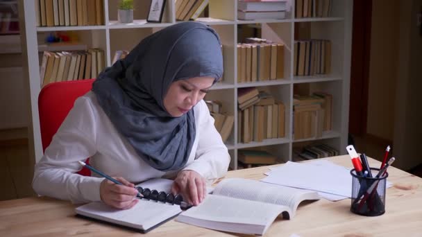 Sesión de primer plano de estudiante musulmana adulta en hiyab estudiando para un examen médico leyendo un libro y tomando notas en el cuaderno de la biblioteca en el interior — Vídeos de Stock