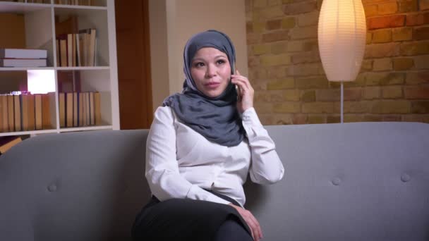 Syuting closeup dari muslim dewasa perempuan di hijab memiliki percakapan telepon sambil duduk di sofa di dalam rumah — Stok Video