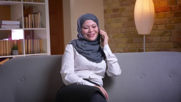 会話を持っているヒジャーブで大人のイスラム教徒の女性のクローズアップシュート携帯電話と陽気に微笑んで自宅で屋内ソファーに座っている間 — ストック動画
