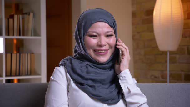 Evde kanepede kapalı otururken bir konuşma telefon gülüyor ve eğlenceli sahip hijab yetişkin Müslüman kadın closeup ateş — Stok video