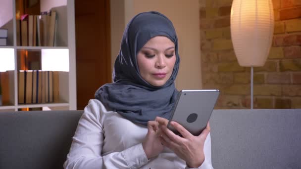 Nahaufnahme einer erwachsenen Muslimin im Hijab mit dem Tablet, während sie zu Hause auf der Couch sitzt — Stockvideo