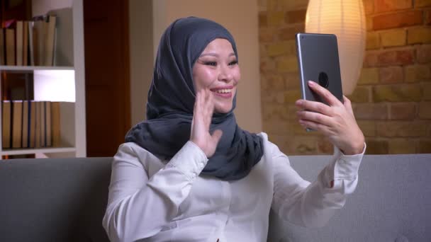 Syuting closeup dari muslim dewasa perempuan di jilbab memiliki panggilan video di tablet sambil duduk di sofa di dalam ruangan di rumah — Stok Video