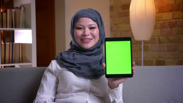 Brote de primer plano de la mujer musulmana adulta en hijab usando la tableta y mostrando la pantalla verde mientras está sentada en el sofá dentro de casa — Vídeo de stock