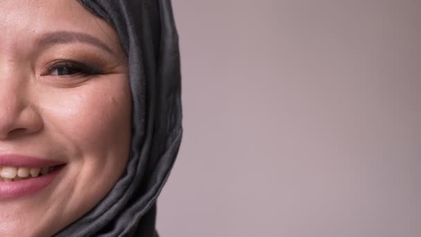 Крупный план съемки полулица взрослой привлекательной мусульманки в хиджабе, смотрящей вперед и счастливо улыбающейся в камеру на изолированном фоне — стоковое видео