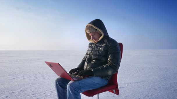 Portret van zakenman in warme jas en kap zitten in de sneeuw woestijn en het werken met laptop vrolijk. — Stockvideo