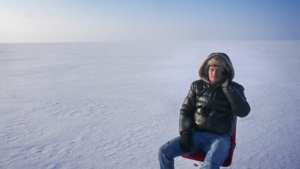 Hombre de negocios alegre en abrigo cálido y capucha en el desierto de nieve girando en la silla y hablando emocionalmente en el teléfono celular . — Vídeos de Stock