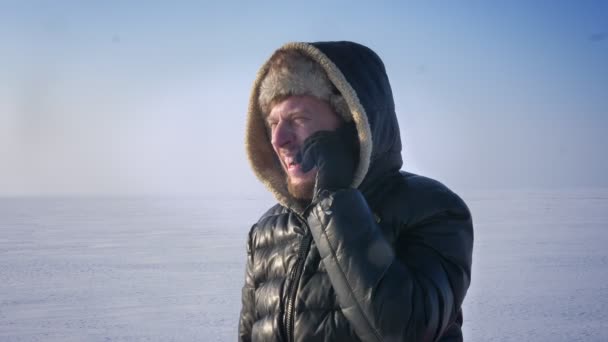 Крупный план портрета бизнесмена в теплом пальто и капюшоне в снежной пустыне разговаривающего по мобильному телефону и смеющегося . — стоковое видео