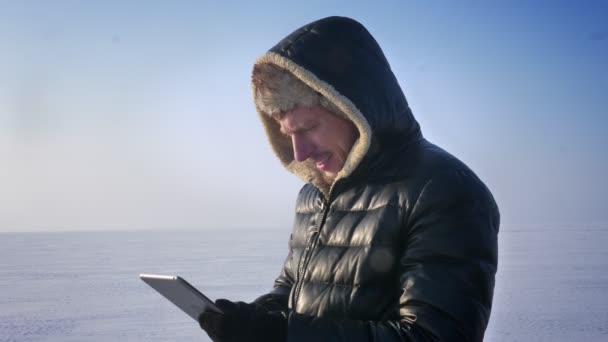 Uomo d'affari in cappotto caldo e cappuccio in piedi sul lago ghiacciato che lavora con tablet si rivolge alla fotocamera e guarda seriamente . — Video Stock