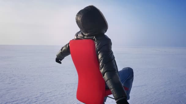 Cheerful infantile businessman in warm coat and hood in snow desert spinning around on red chair. — Stock Video