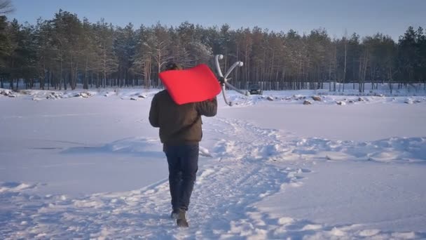 Retrato trasero del hombre de negocios con abrigo cálido y capucha que lleva una silla roja en el hombro sobre el fondo del campo de nieve . — Vídeos de Stock