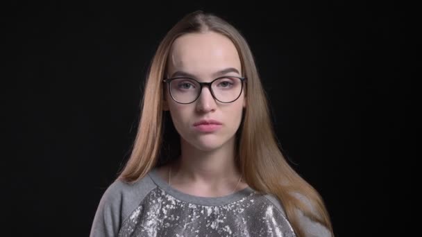 Съемки крупным планом молодой привлекательной хипстерки в очках, смотрящей прямо в камеру на фоне изолированного на черном фоне — стоковое видео